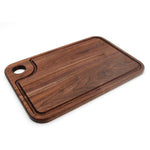 Walnut thumb-hole cutting board with specialty juice groove, angled view on white.