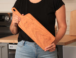 Person holding a cherry narrow paddle board in a kitchen to show its scale and carrying handle.